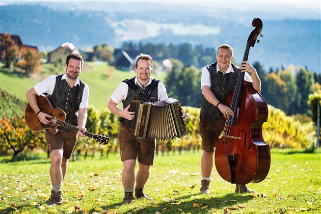 Drei Musiker in traditioneller Tracht stehen in einer malerischen ländlichen Landschaft. Es gibt eine Gitarre, ein Akkordeon und einen Kontrabass.