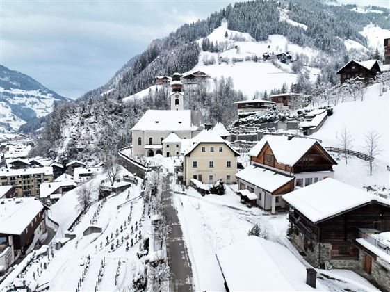 Eine winterliche Landschaft mit schneebedeckten Häusern und Bäumen. Im Hintergrund erheben sich majestätische, verschneite Berge.