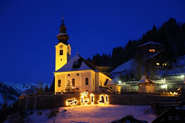 Eine bezaubernde Kirche im Schnee, hell erleuchtet gegen den Nachtblau-Himmel. Im Vordergrund sind Weihnachtsdekorationen mit Figuren zu sehen.