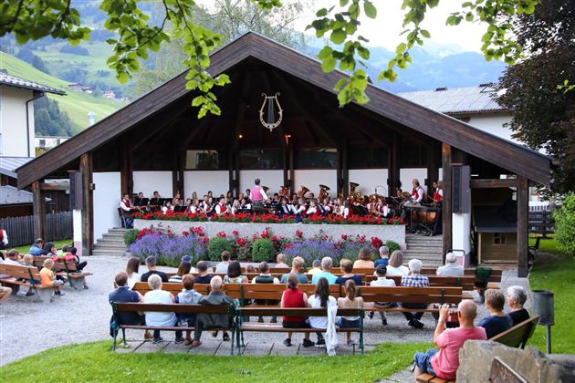 An orchestra is playing outdoors under a pavilion. Many people are sitting on benches and enjoying the music.