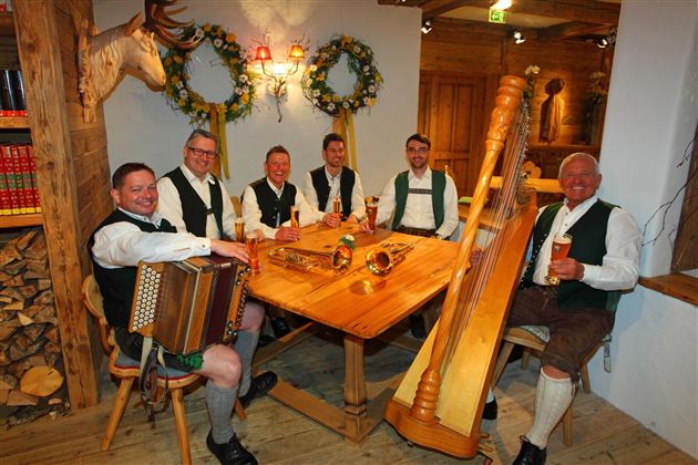 Eine Gruppe von sechs Männern in traditionellen Trachten sitzt an einem Holztisch. Sie spielen Instrumente und genießen Bier in einer gemütlichen, rustikalen Umgebung.