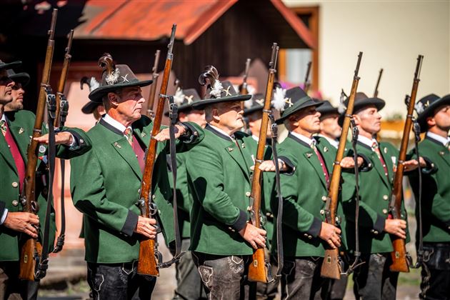 Eine Gruppe von Männern in traditionellen grünen Uniformen steht aufrecht und hält Gewehre in der Hand. Sie scheinen an einer Zeremonie oder Parade teilzunehmen.