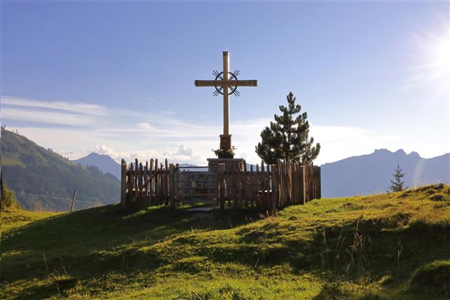 A cross stands on a meadow in the mountains, surrounded by wooden railings and trees. The sky is clear and the landscape appears peaceful.