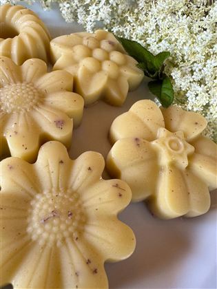 Six handmade soaps in the shape of flowers are on a white plate. In the background, small white blossoms and green foliage can be seen.