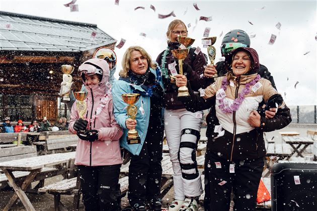 Sieben Frauen feiern den Gewinn von Pokalen in einem festlichen Moment auf einer Skihütte. Im Hintergrund fallen Konfetti und es schneit leicht.