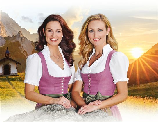 Zwei Frauen in traditionellen Trachten stehen vor einer Berglandschaft. Im Hintergrund sind Berge und ein Sonnenuntergang zu sehen.