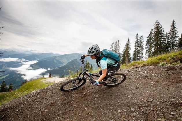 A mountain biker is riding a steep descent among trees. In the background, mountains and clouds can be seen.