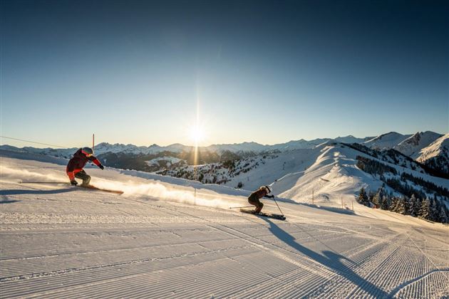 Zwei Skifahrer fahren einen schneebedeckten Hang hinunter. Im Hintergrund sind Berge und ein klarer Himmel bei Sonnenuntergang zu sehen.