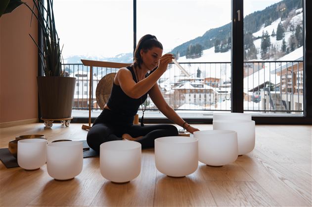 Eine Frau sitzt auf dem Boden und spielt mit Klangschalen in einem hellen Raum. Im Hintergrund sind schneebedeckte Berge und ein Balkon zu sehen.