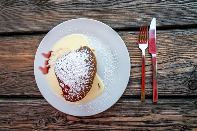 Ein köstliches Dessert auf einem weißen Teller mit Vanillesauce und Puderzucker. Daneben liegen eine Gabel und ein Messer auf einem rustikalen Holztisch.