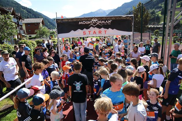 Eine Gruppe von Kindern versammelt sich am Startpunkt eines Wettlaufs. Im Hintergrund sind Berge und eine festliche Atmosphäre zu sehen.
