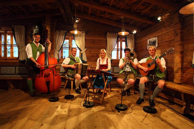 Eine Gruppe von fünf Musikern in traditioneller bayerischer Tracht spielt in einem rustikalen Raum. Sie verwenden verschiedene Instrumente, darunter ein Akkordeon und eine Gitarre.