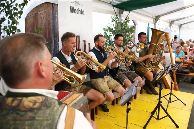A brass band is playing cheerful melodies in a tent. The musicians are wearing traditional costumes and are surrounded by an audience.
