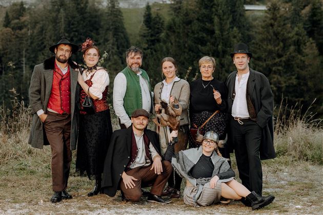 Eine Gruppe von Personen in historischer Kleidung steht im Freien vor einem Wald. Sie posieren freundlich und scheinen an einem besonderen Anlass teilzunehmen.