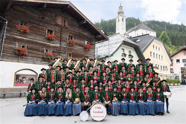 Eine Blaskapelle steht vor traditionellen Gebäuden. Die Musiker tragen festliche Trachten und lächeln in die Kamera.
