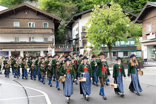 Eine festliche Parade mit Menschen in traditionellen Trachten, die musikalische Instrumente tragen. Im Hintergrund sind typische Holzgebäude und Bäume zu sehen.