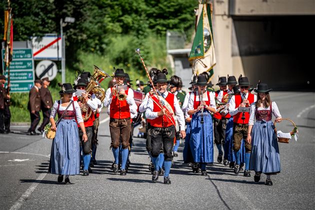 Eine Gruppe von Musikern marschiert auf einer Straße, gekleidet in traditionelle Trachten. Sie spielen verschiedene Instrumente und tragen Fähnchen.