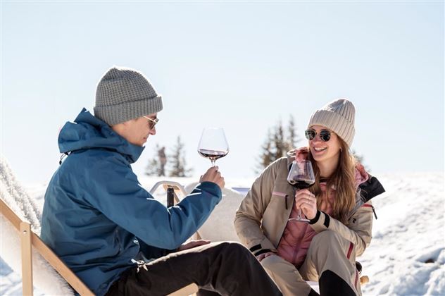 A couple enjoys wine in the wintry landscape. They sit relaxed in the sun, surrounded by snow and fir trees.