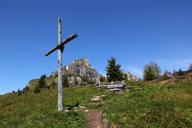 Ein schöner Bergweg mit einem Holzkreuz und einer Sitzgelegenheit. Im Hintergrund sind majestätische Berge und ein klarer blauer Himmel zu sehen.