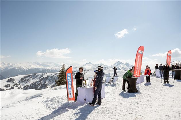 A sunny winter landscape with snow and mountains in the background. People are enjoying an event with standing tables and brand flags at the summit.