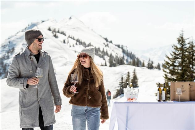 A couple enjoys a sunny day in the mountains, holding wine glasses in their hands. In the background, there are snow-covered mountains and a table with wine bottles.