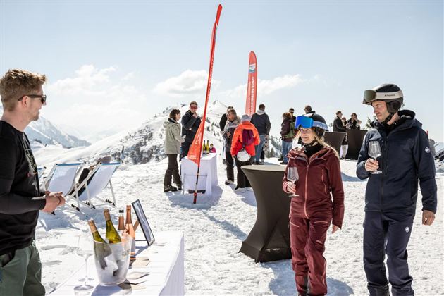 A group of people is enjoying a wine tasting in the mountains. The scene takes place on snow, surrounded by a breathtaking mountain landscape.