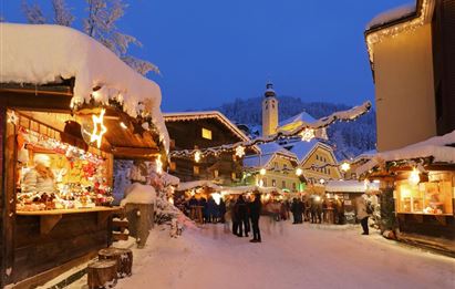 Eine winterliche Marktszene mit schneebedeckten Holzhütten und festlicher Beleuchtung. Menschen genießen die Atmosphäre und die winterlichen Stände.