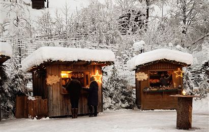 Eine winterliche Landschaft mit schneebedeckten Hütten. Menschen stehen an den Hütten und genießen die festliche Atmosphäre.