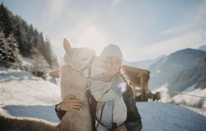 A woman hugs a llama in a snow-covered landscape. In the background, mountains and a wooden house are visible.