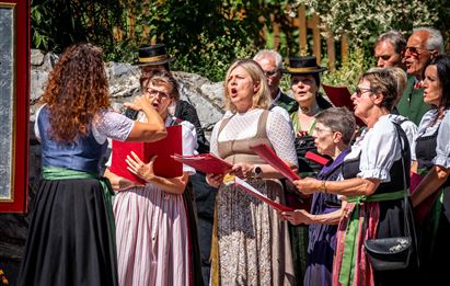 Eine Gruppe von Sängerinnen und Sängern tritt im Freien auf. Sie tragen traditionelle Kleidung und singen mit Freude.