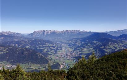 Ein beeindruckender Blick auf ein Tal mit grünen Wiesen und einem kleinen Dorf. Im Hintergrund befinden sich majestätische Berge unter einem klaren Himmel.
