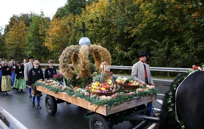 Eine festliche Parade mit einem Wagen, der mit Obst und Erntegaben geschmückt ist. Menschen in traditionellen Trachten begleiten den Wagen entlang der Straße.