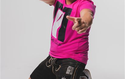 Ein männlicher Model sitzt kniend und trägt ein pinkes T-Shirt. Er trägt Sonnenbrillen und posiert enthusiastisch mit einem Lächeln.