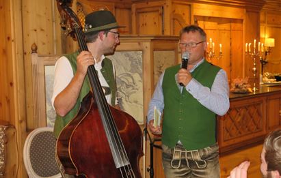 Zwei Männer in traditioneller Tracht stehen in einem gemütlichen Raum. Einer spielt Kontrabass, während der andere ein Mikrofon hält und spricht.