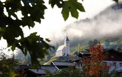 A picturesque church stands on a hill, surrounded by green forests and mist. Autumn leaves frame the scene and add a burst of color.