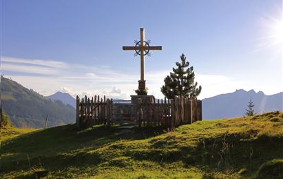 Ein Kreuz steht auf einer Wiese in den Bergen, umgeben von Holzgeländer und Bäumen. Der Himmel ist klar und die Landschaft wirkt friedlich.