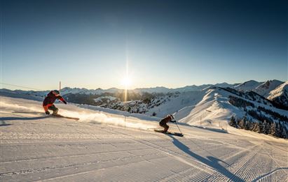 Zwei Skifahrer fahren einen schneebedeckten Hang hinunter. Im Hintergrund sind Berge und ein klarer Himmel bei Sonnenuntergang zu sehen.