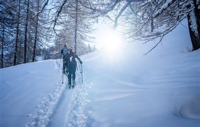Zwei Personen wandern durch eine verschneite Landschaft. Die Sonne scheint und sorgt für eine helle, winterliche Atmosphäre.