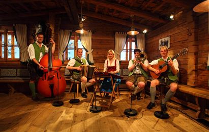 Eine Gruppe von fünf Musikern in traditioneller bayerischer Tracht spielt in einem rustikalen Raum. Sie verwenden verschiedene Instrumente, darunter ein Akkordeon und eine Gitarre.