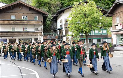Eine festliche Parade mit Menschen in traditionellen Trachten, die musikalische Instrumente tragen. Im Hintergrund sind typische Holzgebäude und Bäume zu sehen.