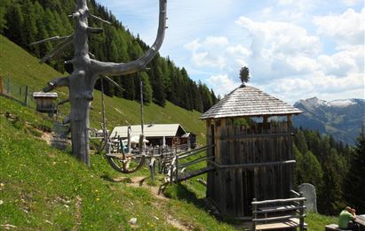 Ein malerischer Berglandschaft mit einer Holzstruktur und einem Aussichtsturm. Im Hintergrund sind grüne Wiesen und schneebedeckte Berge zu sehen.