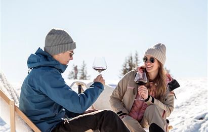 A couple enjoys wine in the wintry landscape. They sit relaxed in the sun, surrounded by snow and fir trees.