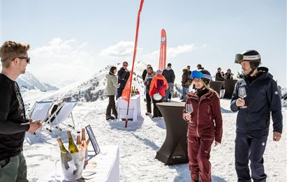 Eine Gruppe von Menschen genießt eine Weindegustation in den Bergen. Die Szene findet auf Schnee statt, umgeben von einer atemberaubenden Berglandschaft.