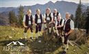 Eine Gruppe von sechs Männern in traditioneller bayerischer Tracht steht auf einem Berg. Im Hintergrund sind beeindruckende Alpenlandschaften zu sehen.