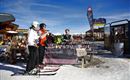 A group of skiers is standing on a snowy area and talking to a seller. In the background, ski buildings and a clear mountain landscape can be seen.