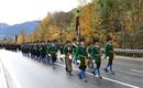 A parade with uniformed participants marching in colorful costumes. In the background, autumn trees and mountains can be seen.