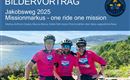 Drei Radfahrer tragen pinke Trikots und lächeln in die Kamera. Im Hintergrund ist eine schöne Landschaft mit Bergen und Wasser zu sehen.