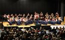 Ein Blasorchester spielt auf einer Bühne vor einem begeisterten Publikum. Die Musiker tragen traditionelle Kleidung und stehen in einer Reihe.