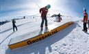Eine Person fährt auf Skiern über ein Snowboard-Rail in einer verschneiten Berglandschaft. Im Hintergrund sind weitere Skifahrer und eine wunderschöne alpine Kulisse zu sehen.