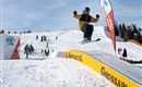 Ein Snowboarder springt über eine Rampe im Skigebiet Grossarltal. Im Hintergrund sind weitere Skifahrer und Skilehrer sichtbar.